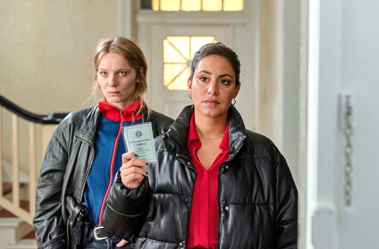 Zwei Frauen stehen in einem Eingangsbereich. Die vordere Frau hält einen Polizeiausweis in der Hand. Sie trägt eine schwarze Steppjacke und ein rotes Hemd und blickt ernst. Die hintere Frau, in einer Lederjacke und einem roten Schal, schaut aufmerksam in dieselbe Richtung. Im Hintergrund befinden sich eine Treppe und eine Tür mit Glasfenstern.