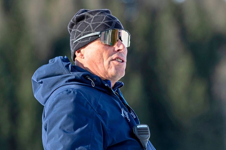 Ein Mann in Winterbekleidung mit Sonnenbrille und Mütze steht im Freien. An seiner Jacke hängt ein Funkgerät. Hintergrund ist unscharf mit Bäumen.