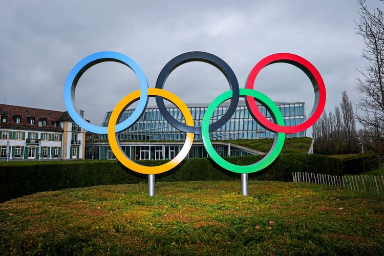 Die olympischen Ringe stehen auf einer Grünfläche vor dem Gebäude des Internationalen Olympischen Komitees (IOC) im olympischen Haus in Lausanne, Schweiz, unter einem bewölkten Himmel.