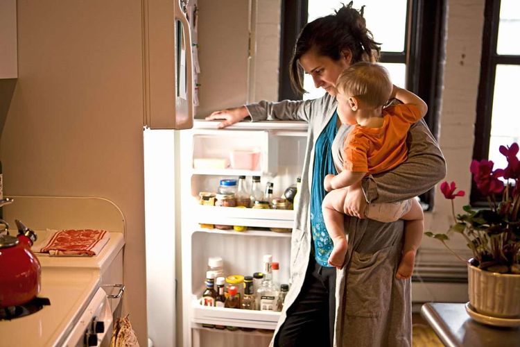 Eine Frau steht mit ihrem Kleinkind am Arm vor dem Kühlschrank.