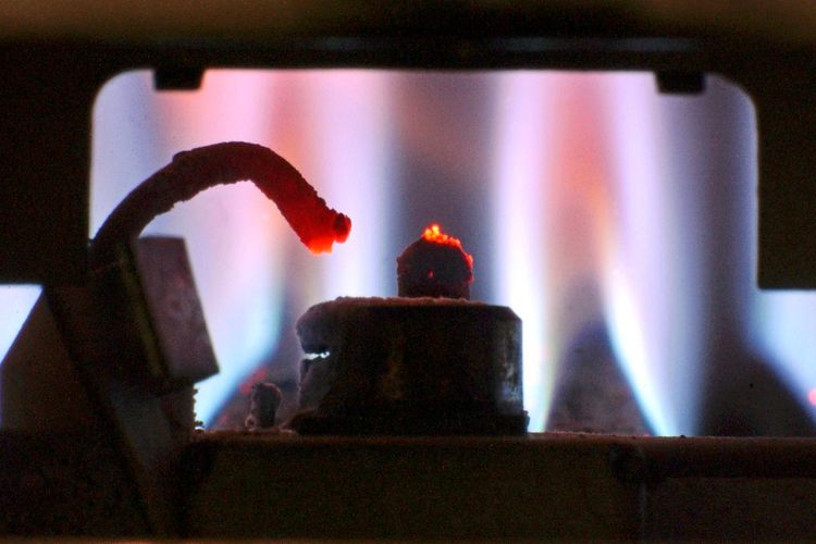 Zündvorrichtung einer Gastherme mit leuchtender Flamme in einem Haushalt, im Hintergrund blaue Gasflammen sichtbar.