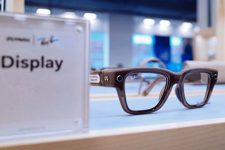 Eine Nahaufnahme einer braunen Brille von Ray-Ban in Zusammenarbeit mit Meta, ausgestellt in einem Elektronikgeschäft. Im Hintergrund sind verschwommene Lichter und Regale zu sehen, während der Fokus auf der Brille liegt. Ein Schild mit der Aufschrift „Display“ ist links sichtbar.