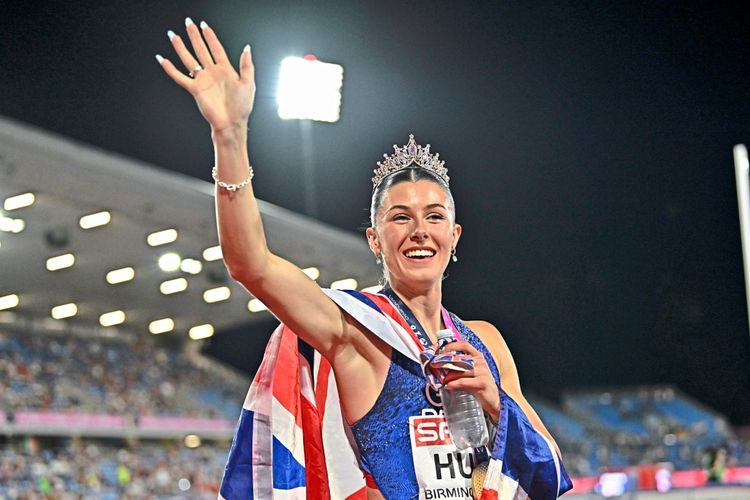 Eine Frau in Sportkleidung mit einer Krone auf dem Kopf winkt und lächelt vor einem beleuchteten Stadion. Sie trägt eine Flagge über den Schultern und eine Medaille um den Hals.