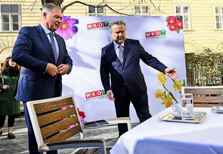 Zwei Männer in Anzügen stehen im Freien vor einem Banner der Wirtschaftskammer Wien (WKW) mit Blumenmotiven. Einer der Männer deutet mit der Hand auf einen Tisch im Vordergrund, auf dem ein Tablett mit Gläsern und einer Kaffeetasse steht. Im Hintergrund sind eine Frau, Büsche und ein Gebäude mit Fenstern zu sehen.