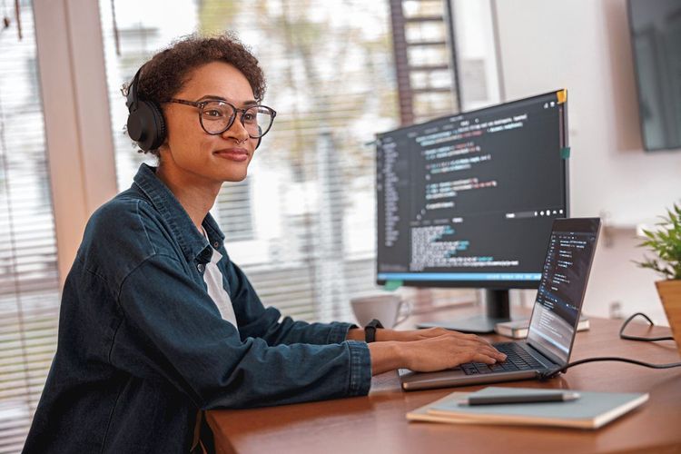 Junge Frau mit Kopfhörern sitzt vor einem Laptop und Bildschirm und schreibt Code
