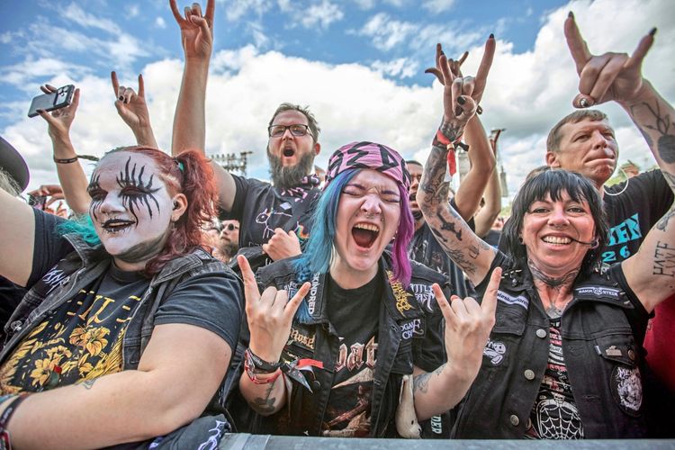 Eine Gruppe von begeisterten Metal-Fans steht in der Menge des Wacken Open Air Festivals. Sie tragen Band-T-Shirts, Kutten und Make-up, heben die Hände in die Luft und zeigen das typische 
