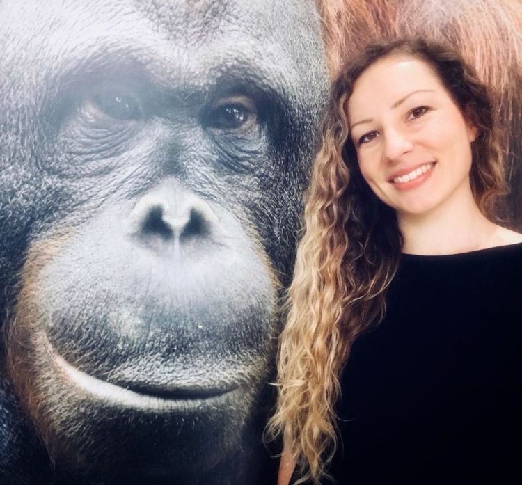 Eine junge Frau mit langen, gewellten, braun-blonden Haaren und einem schwarzen Pullover steht neben dem überdimensionalen Bild eines Orang-Utans.