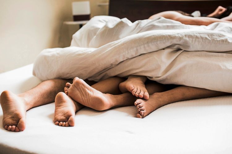 Drei Paar Füße ragen unter einer weißen Bettdecke hervor, liegend auf einem Bett in einem hellen Schlafzimmer.