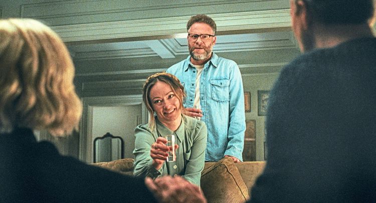 Eine Frau sitzt lächelnd auf einem Sofa und hält ein Glas in der Hand. Hinter ihr steht ein Mann mit Brille und Jeanshemd. Im Vordergrund sind unscharf zwei Personen von hinten zu sehen. Die Szene spielt in einem Wohnzimmer mit heller Beleuchtung.