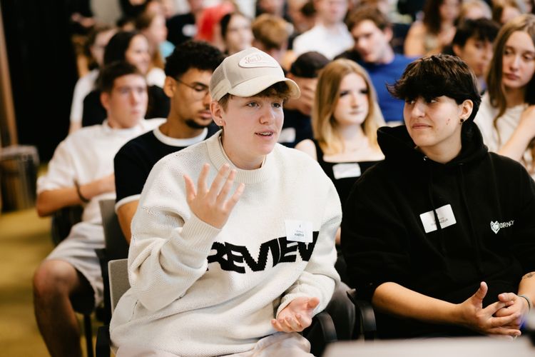 Eine Gruppe junger Menschen sitzt in einem Veranstaltungsraum. Im Vordergrund sprechen zwei Personen miteinander, eine trägt einen weißen Pullover und eine Kappe, die andere einen schwarzen Hoodie. Im Hintergrund sind weitere Teilnehmende zu sehen.