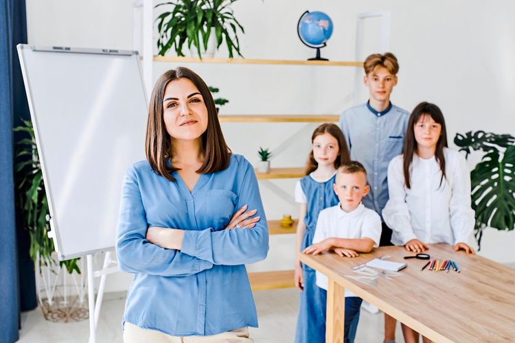 Eine Frau in einer blauen Bluse steht mit verschränkten Armen lächelnd im Vordergrund eines Klassenzimmers. Im Hintergrund stehen vier Kinder unterschiedlichen Alters vor einem Tisch mit Schreibmaterialien. Im Raum befinden sich ein Whiteboard, Regale, Pflanzen und ein Globus.