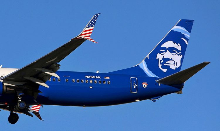 Ein Alaska Airlines Boeing 737-900ER Flugzeug mit einer dunkelblauen Lackierung und einem Gesichtsmotiv auf dem Heck, sowie einer Heckflosse mit einem Muster der US-Flagge, im Landeanflug unter klarem Himmel.