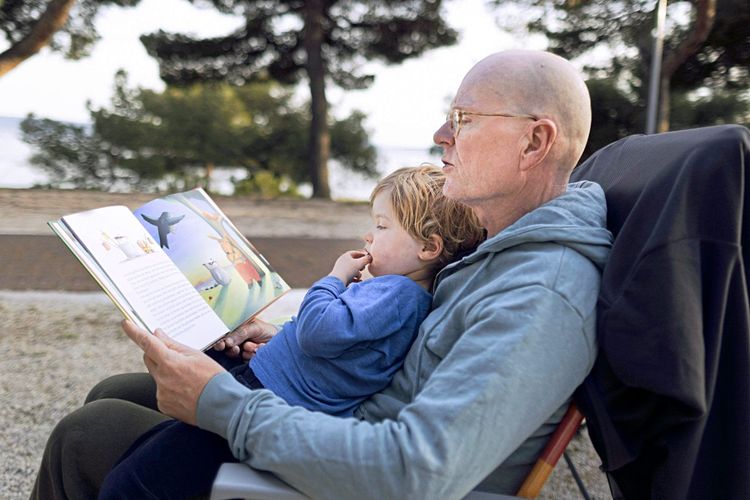 Ein Großvater sitzt mit seinem Enkel auf dem Schoß in einem Sessel und liest ihm aus einem Buch vor, in das beide schauen.