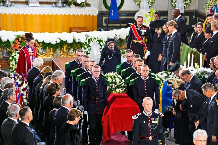 Ein Sarg wird von uniformierten Trägern aus einer Kirche getragen, umgeben von trauernden Personen in festlicher Kleidung. Im Hintergrund ist ein mit Blumen geschmückter Altar zu sehen.