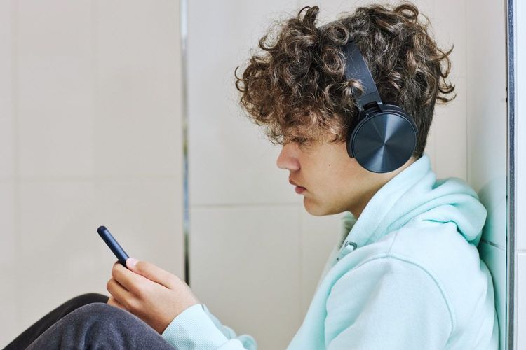 Junge sitzt an der Wand, Handy in der Hand mit Kopfhörern.
