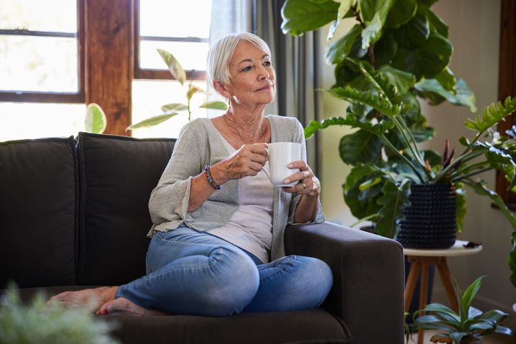 Eine ältere Frau sitzt mit Kaffeehäferl in der Hand auf einem Sofa.