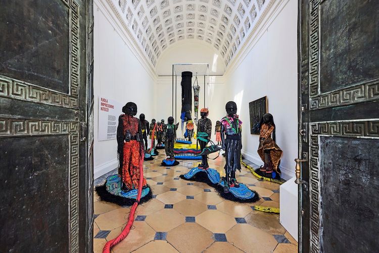 Blick durch geöffnete verzierte Türen in einen Ausstellungsraum mit farbenfrohen Skulpturen menschlicher Figuren. Der Raum hat eine hohe, gewölbte Decke und helle Wände. Schilder und Kunstwerke sind an den Wänden angebracht.