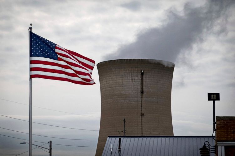Eine US-amerikanische Flagge weht im Vordergrund, während im Hintergrund ein Kühlturm eines Kohle- und Erdgaskraftwerks der Northern Indiana Public Service Company (NIPSCO) in Michigan City, Indiana, emporragt. Aus dem Turm entweicht Rauch in einen bewölkten Himmel.
