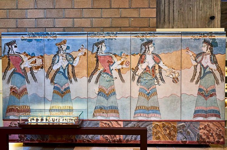 Wandgemälde mit rekonstruierter minoischer Fresko-Darstellung aus der Antike. Mehrere Frauen in detailreichen, bunten Gewändern mit lockigem Haar sind abgebildet. Im Vordergrund eine Glasvitrine mit kleinen Artefakten. Hintergrund aus Ziegel- und Betonwand.