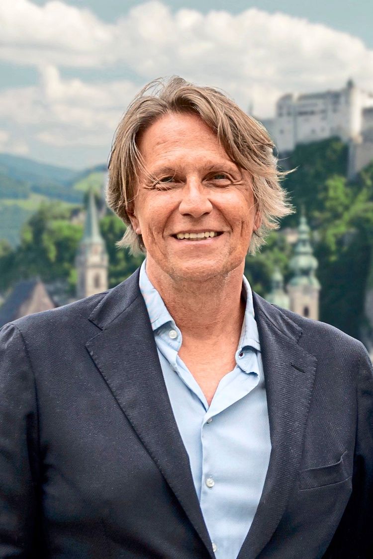 Ein lächelnder Mann mittleren Alters steht vor einer malerischen Kulisse mit Bergen, einer Kirche und einer Burg im Hintergrund. Er trägt ein dunkelblaues Sakko und ein hellblaues Hemd.