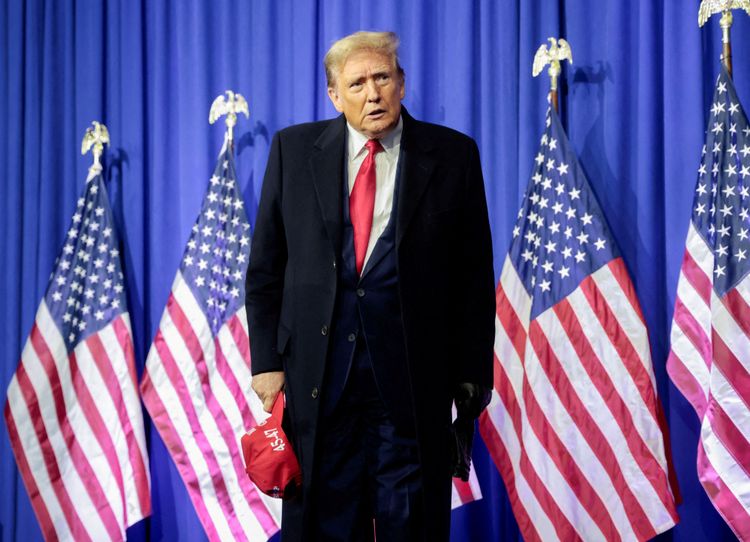 Donald Trump im Mantel mit roter Krawatte und roter Kappe in der rechten Hand; US-Flaggen im Hintergrund
