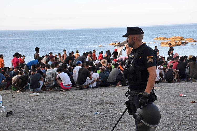 Ein Polizist in Uniform steht am Strand, während im Hintergrund eine große Gruppe von Menschen in der Nähe des Meeres sitzt. Die Szene zeigt eine Versammlung von Migranten, die medizinische Versorgung und Hilfe von Freiwilligen und Fachleuten erhalten. Im Hintergrund sind Felsen im Wasser zu sehen.