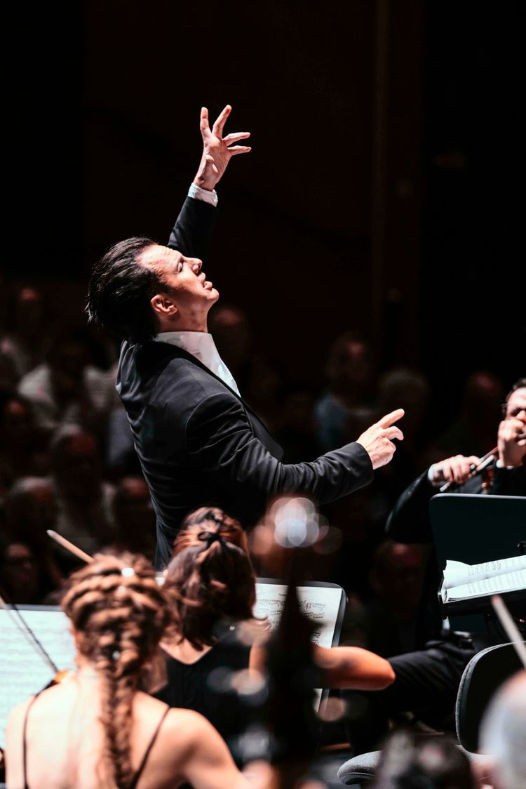 Ein Dirigent in einem schwarzen Anzug dirigiert mit ausdrucksvollen Bewegungen ein Orchester während eines Konzerts. Im Vordergrund sind unscharf Orchestermusiker und Noten sichtbar.