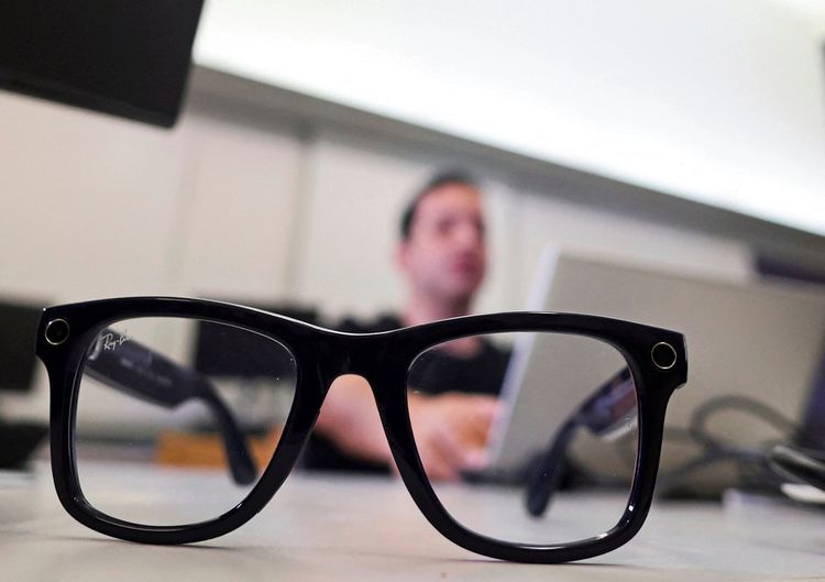 Eine schwarze Brille mit integrierter Technologie liegt im Vordergrund auf einem Tisch, während eine Person unscharf im Hintergrund an einem Laptop arbeitet.