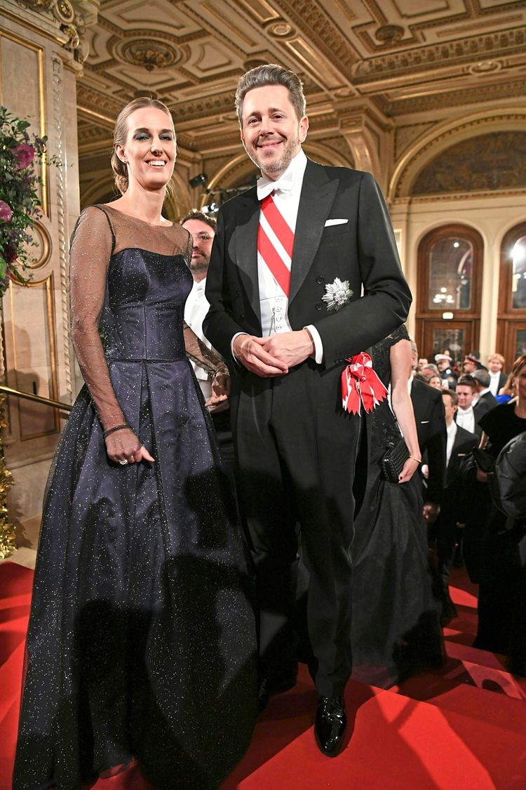 Ein Mann und eine Frau stehen lächelnd auf einem roten Teppich in einem prunkvollen Saal, vermutlich während des Wiener Opernballs. Der Mann trägt einen Anzug mit Orden, die Frau ein langes glitzerndes Abendkleid. Im Hintergrund sind weitere festlich gekleidete Personen und ein reich verziertes Interieur zu sehen.