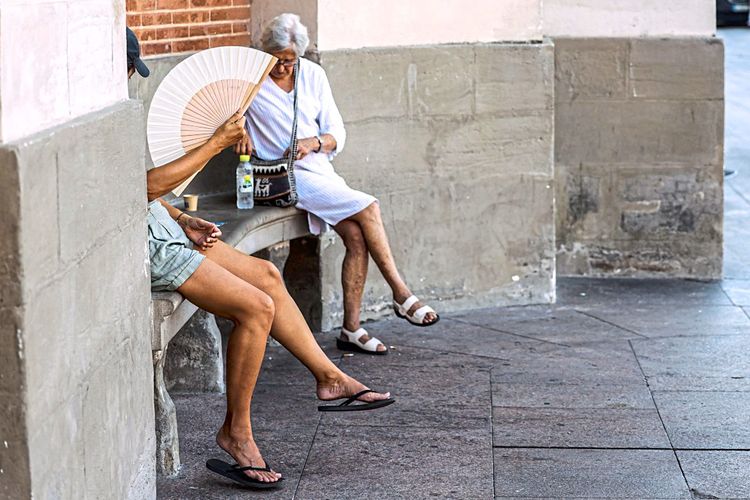 Zwei Frauen sitzen auf einer Bank in einer Ecke auf den Straßen von Toulouse am 23. August 2023: Eine Frau hält einen Fächer, die andere hat eine Wasserflasche neben sich stehen und kramt in ihrer Tasche.