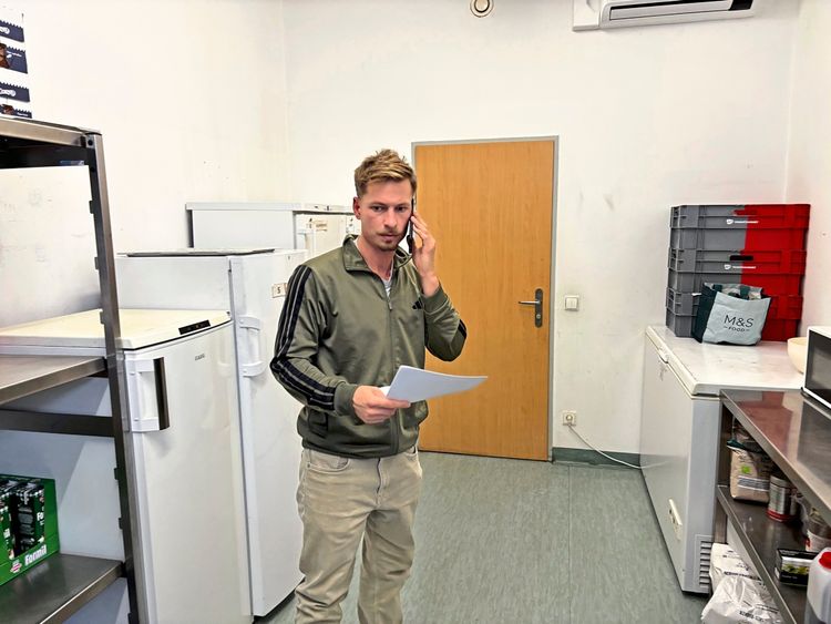 Ein Mann steht in einem Kühlraum und spricht am Telefon, während er ein Blatt Papier in der Hand hält. Im Hintergrund sind Kühlschränke, Regale und Kisten zu sehen.