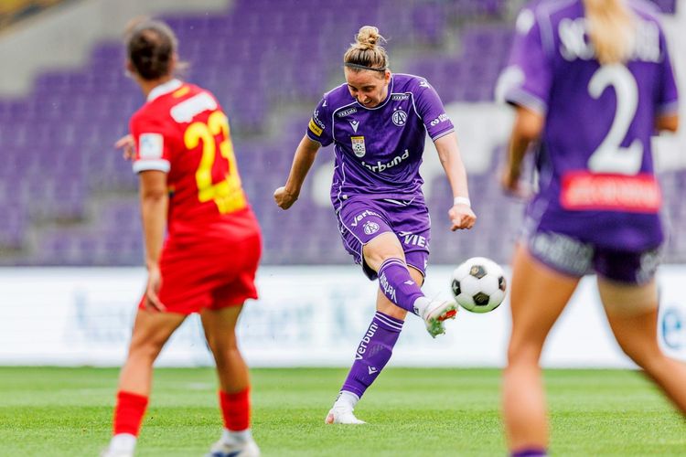 Eine Fußballspielerin des FK Austria Wien Frauen im lila Trikot schießt den Ball während eines Spiels der ADMIRAL Frauen Bundesliga gegen SKN St. Pölten Frauen. Im Hintergrund sind Spielerinnen beider Teams und leere Tribünen zu sehen.