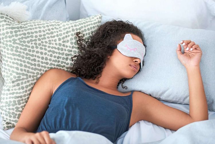 Junge Frau mit dunklen, lockigen Haaren liegt im Bett und schläft. Sie trägt eine Schlafmaske.