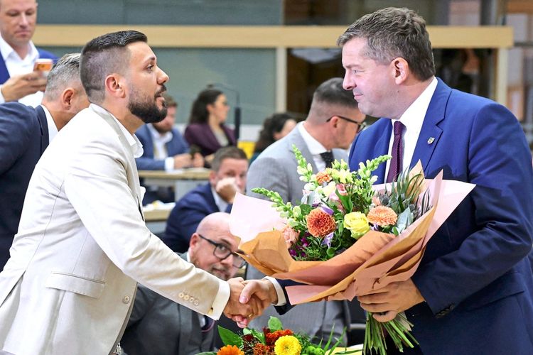 Ein Mann in einem weißen Anzug schüttelt die Hand eines Mannes in einem blauen Anzug und überreicht ihm einen Blumenstrauß. Im Hintergrund sitzen mehrere Personen in einem Sitzungsraum.