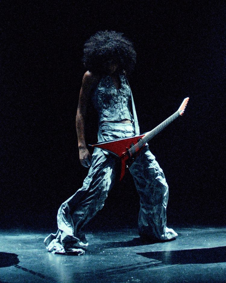Eine Person mit voluminösem, lockigem Haar steht auf einer dunklen Bühne und spielt eine rote E-Gitarre. Sie trägt ein glänzendes, texturiertes Outfit mit weiter Hose. Die Szene ist dramatisch beleuchtet.