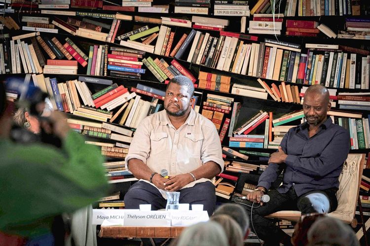 Zwei Männer sitzen während einer Diskussion vor einem Bücherregal. Der Mann in der Mitte trägt ein beiges Hemd und hält ein Mikrofon in der Hand, während der Mann rechts ein dunkles Hemd trägt. Im Hintergrund sind zahlreiche Bücher auf schrägen Regalen sichtbar. Es handelt sich um eine Veranstaltung im Rahmen eines Literaturfestivals.