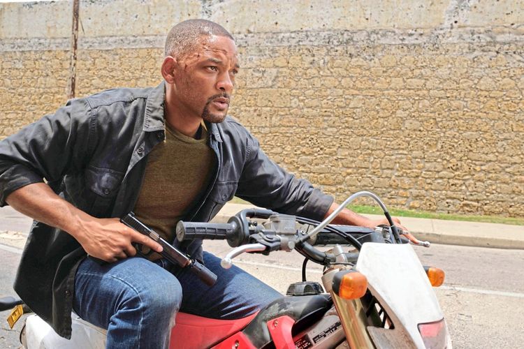 Eine Actionszene zeigt einen Mann auf einem roten Motorrad. Er trägt eine dunkle Jacke und ein grünes T-Shirt, während er angespannt schaut. In seiner rechten Hand hält er eine Pistole. Im Hintergrund ist eine Steinmauer und eine Straße zu sehen.