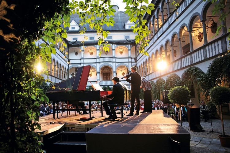 Ein Konzert im Innenhof von Schloss Greinburg während der Donaufestwochen im Strudengau. Musiker spielen auf einer beleuchteten Bühne vor einem Publikum, umgeben von historischer Architektur und Pflanzen.