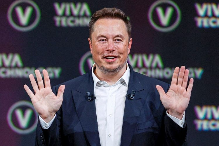 Elon Musk im Porträt in Anzug und weißem Hemd, er hält die Hände hoch, sodass man seine Handflächen sieht. 