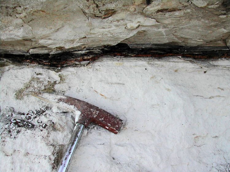 Heller Fels, durchzogen von einer dunklen Schicht. Davor ein spitzer Geologenhammer.