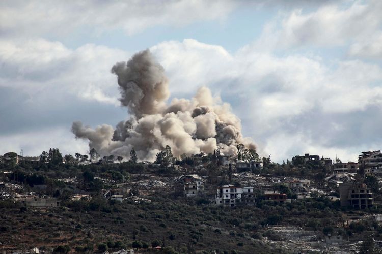Dichter Rauch steigt von einer Explosion in einer hügeligen Landschaft auf, umgeben von vereinzelten Gebäuden und Vegetation unter einem bewölkten Himmel.