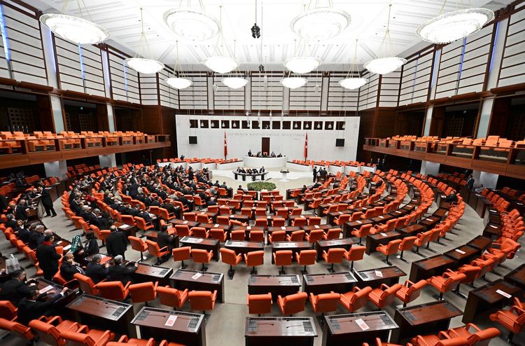 Innenansicht des türkischen Parlaments (Große Nationalversammlung der Türkei) während einer Sitzung. Der Versammlungssaal ist in einem halbrunden Stil gestaltet, mit orangefarbenen Sitzen, einer erhöhten Bühne und hellen Kronleuchtern an der Decke. Menschen sitzen oder stehen im Saal, während Diskussionen stattfinden. Türkische Fahnen sind sichtbar.