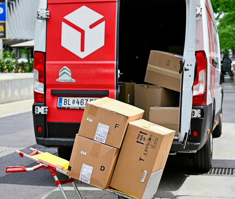 Ein Lieferwagen mit geöffneter Tür, aus dem mehrere Pakete auf einem Transportwagen entladen werden. Aufgenommen in einer städtischen Umgebung in Wien.