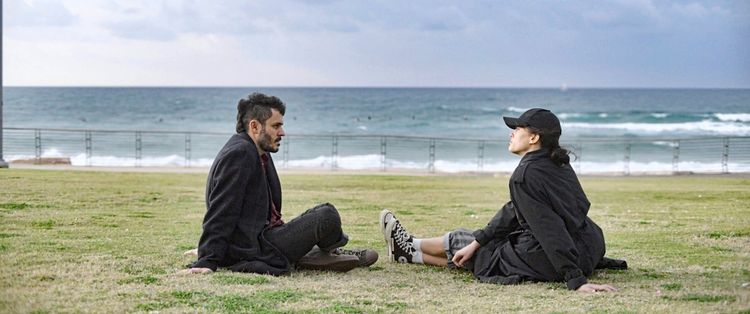 Zwei Personen sitzen auf einer Wiese und unterhalten sich, mit Blick auf das Meer im Hintergrund. Es ist bewölkt, und eine Metallabsperrung trennt die Wiese vom Strand.