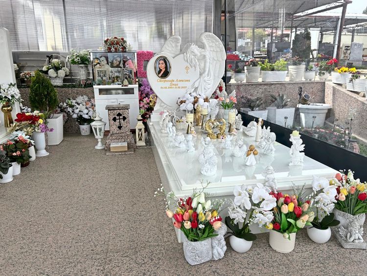 Ein geschmücktes Grab mit weißem Grabstein in Herzform, verziert mit Engelsflügeln und einem Bild einer Frau. Der Grabstein ist umgeben von zahlreichen Blumensträußen, Statuen von Engeln und anderen dekorativen Elementen in Weiß und Gold. Im Hintergrund befinden sich weitere Blumenarrangements und Blumenkästen.