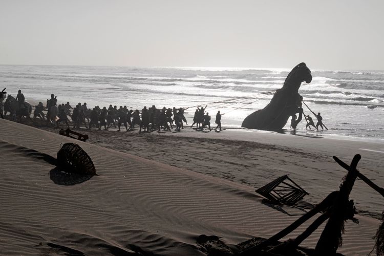Eine große Gruppe von Menschen zieht mit Seilen eine riesige Pferdeskulptur aus dem Meer an einen Sandstrand. Im Vordergrund liegen verstreute Gegenstände im Sand, während Wellen im Hintergrund brechen.