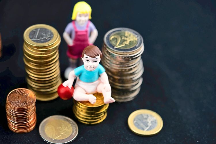 Zwei Spielfiguren, ein Kind und ein Baby, sitzen zwischen gestapelten Euro-Münzen auf schwarzem Hintergrund.