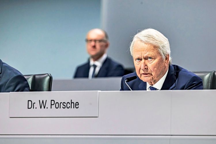 Ein älterer Mann mit weißem Haar sitzt an einem Tisch mit einem Namensschild 