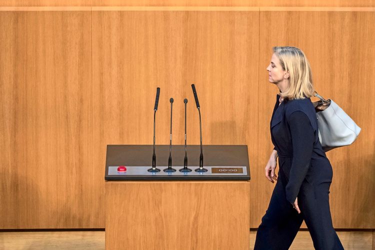 Eine Frau in dunkler Kleidung mit einer hellen Tasche über der Schulter geht an einem Rednerpult mit Mikrofonen vorbei. Im Hintergrund ist eine holzvertäfelte Wand zu sehen. Szene aus einer Sondersitzung des österreichischen Nationalrats.
