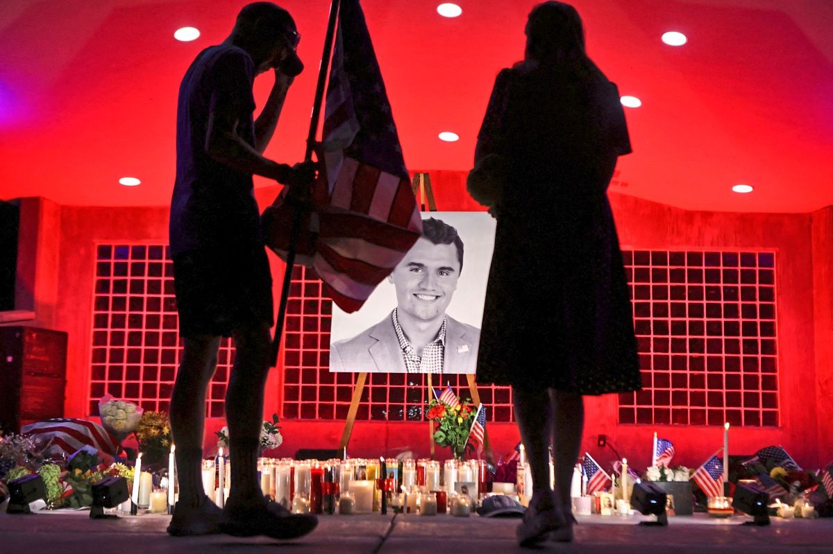 Two people stand in a red-lit area in front of a memorial site for Charlie Kirk with candles, flowers, small US flags and a large portrait.